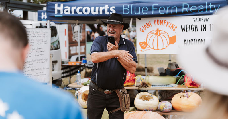 Exhibitor in a hat beside giant pumpkins at the Harcourts Blue Fern Realty site